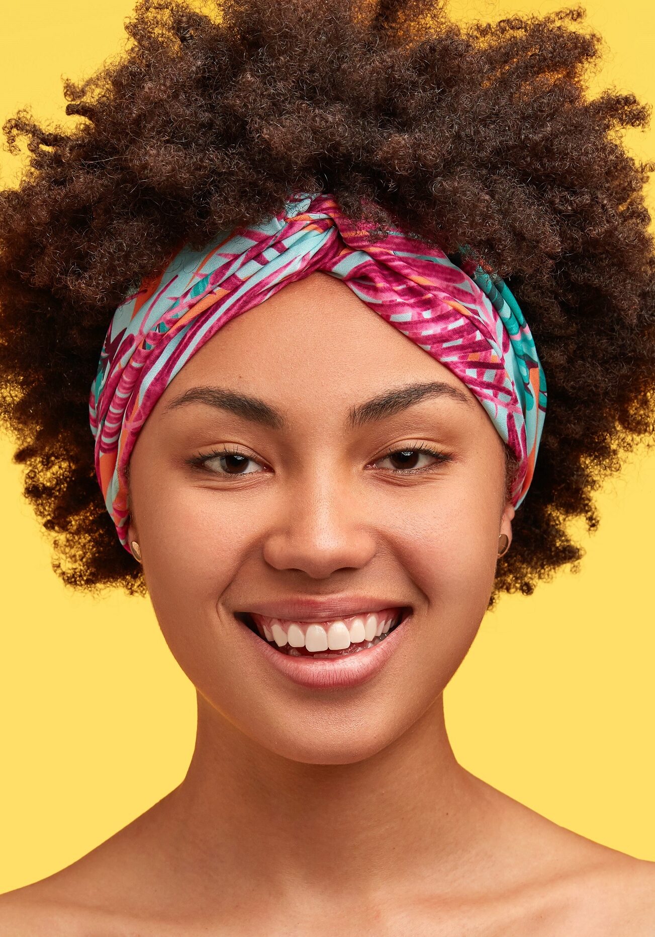 Glad lovely dark skinned female with toothy smile, dressed in striped t shirt, headband, expresses positiveness, isolated over yellow background. Cheerful African American teenager poses indoor